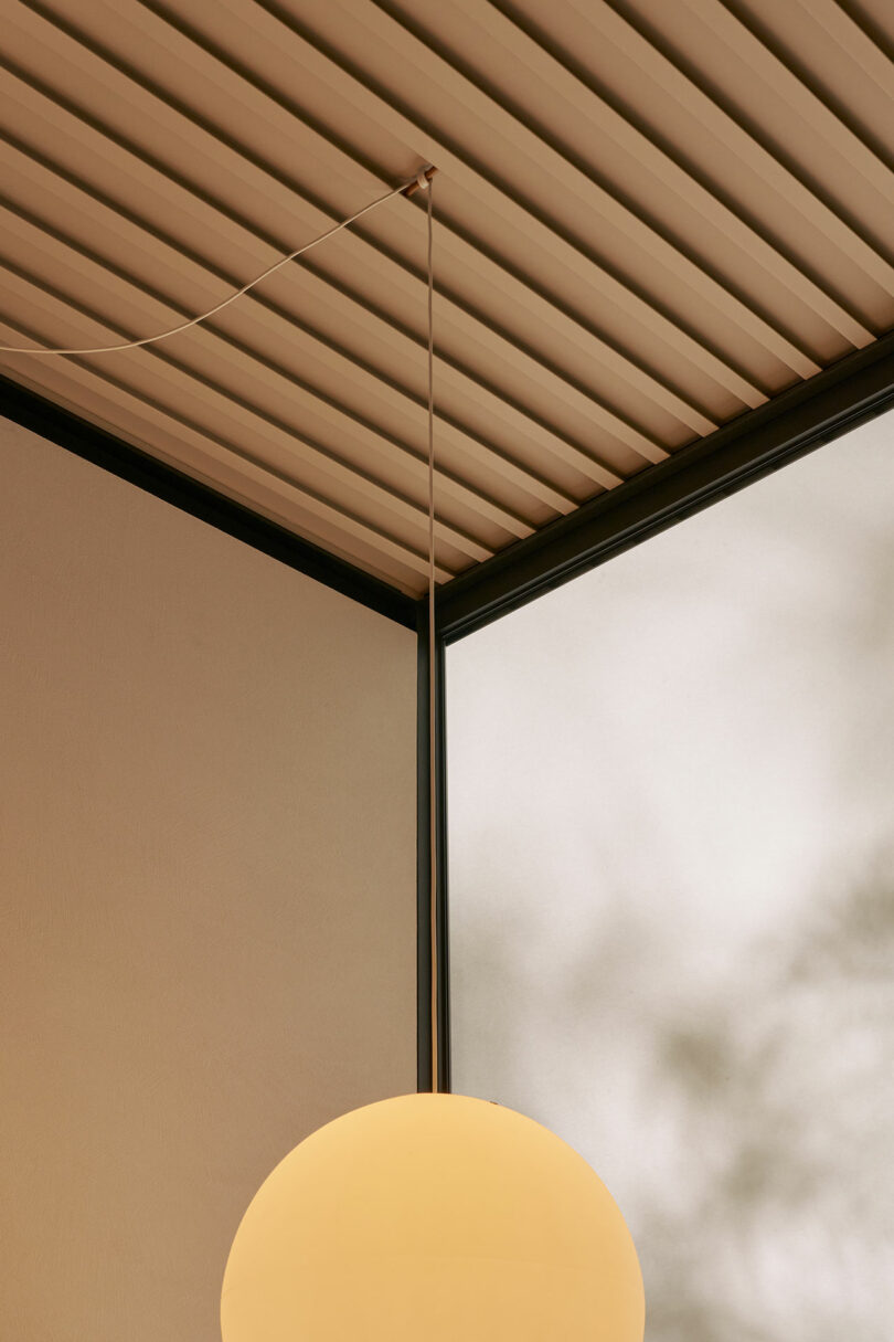 A round pendant light hangs from a ceiling with parallel beams near the corner of a room with frosted glass windows and soft, diffused natural light.