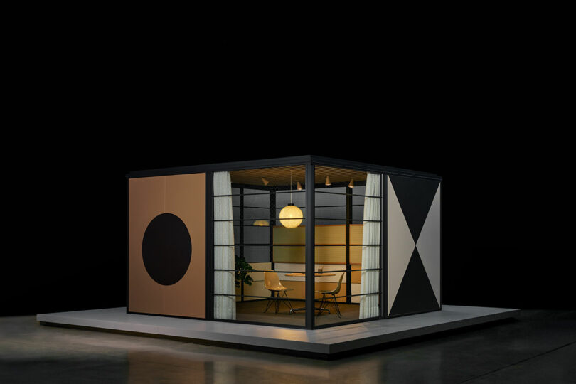 A modern, glass-walled meeting room with geometric-patterned exterior panels stands on a platform, furnished with a round table, four chairs, and a hanging circular light.