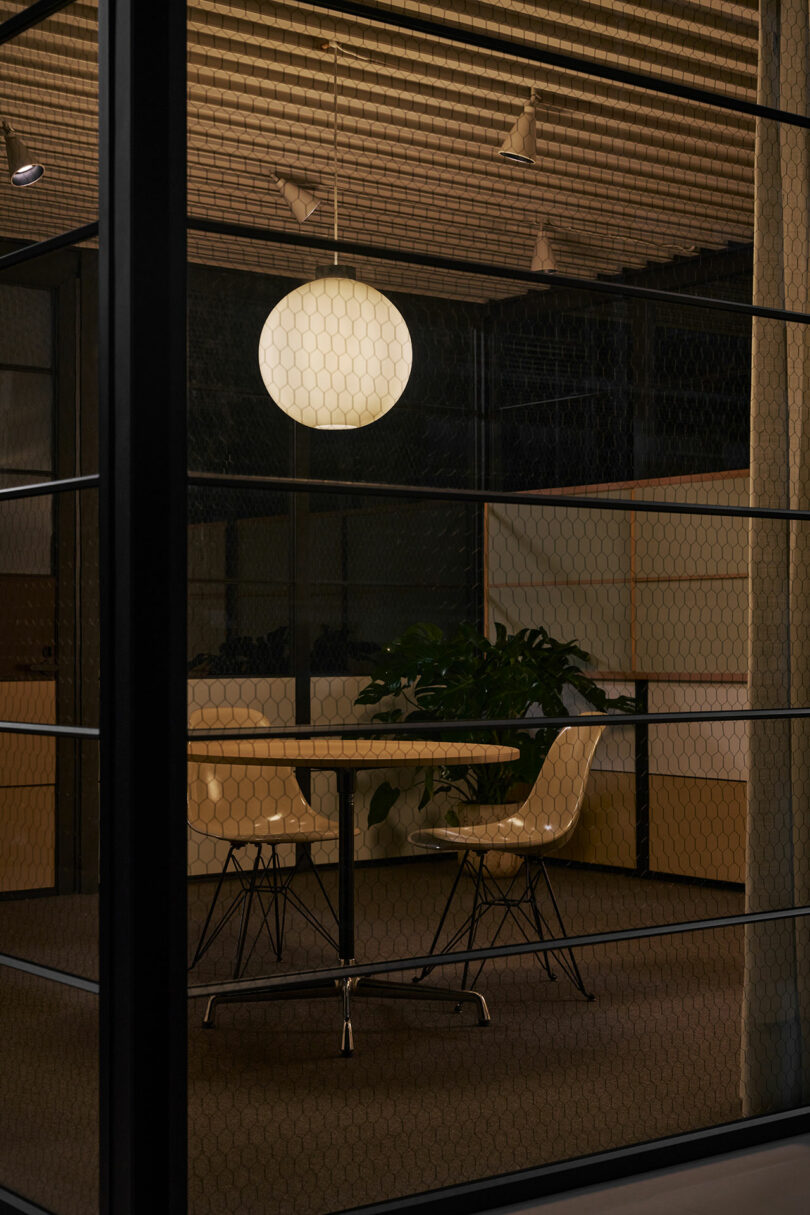 A modern meeting room with a round table, two white chairs, a large round ceiling light, and a potted plant, viewed through glass walls.
