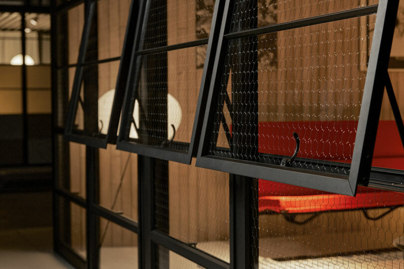 Close-up view of black-framed, open windows with wire mesh, revealing a red bench and a white lamp inside a modern interior space.