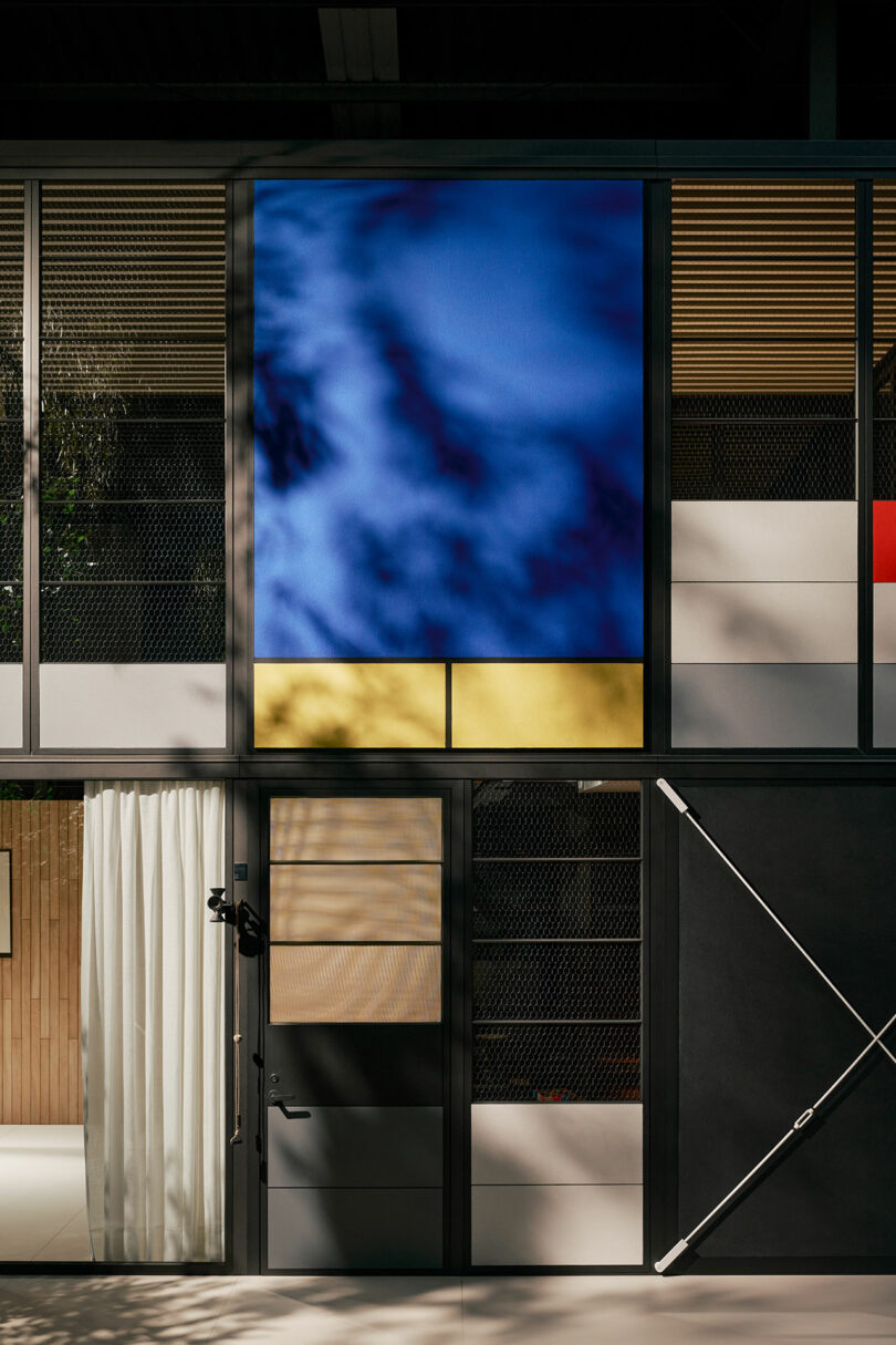 Modern building facade with a large blue abstract panel above a yellow section, featuring glass, metal, and wood elements, along with a black door and mesh details.