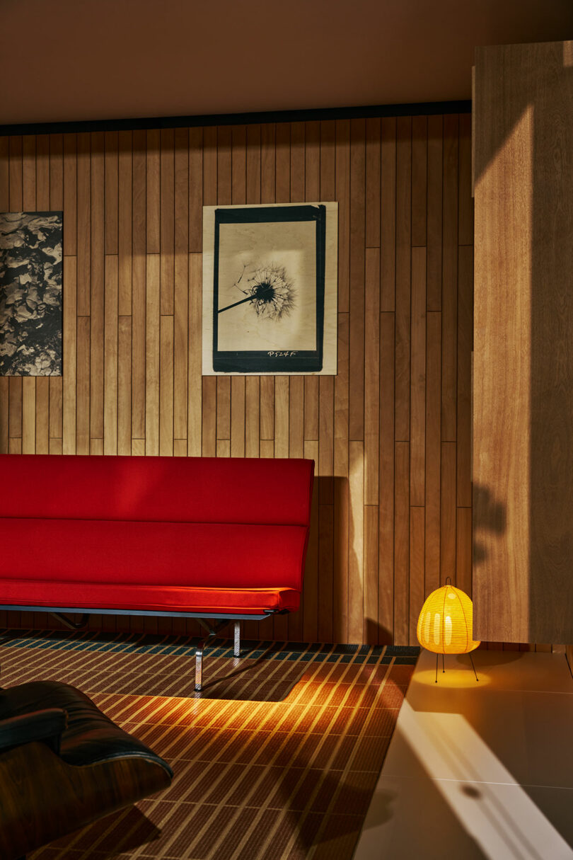 A mid-century modern room with wood panel walls, a red sofa, two framed artworks, a striped rug, and a small yellow lamp on the floor.