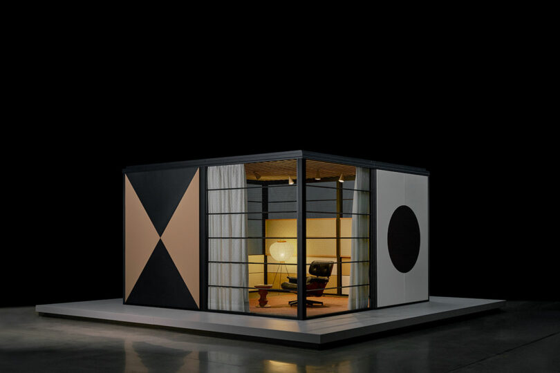 A modern, box-shaped room with geometric patterns on its exterior walls, featuring large windows, a chair, a side table, and a lamp inside, set against a black background.