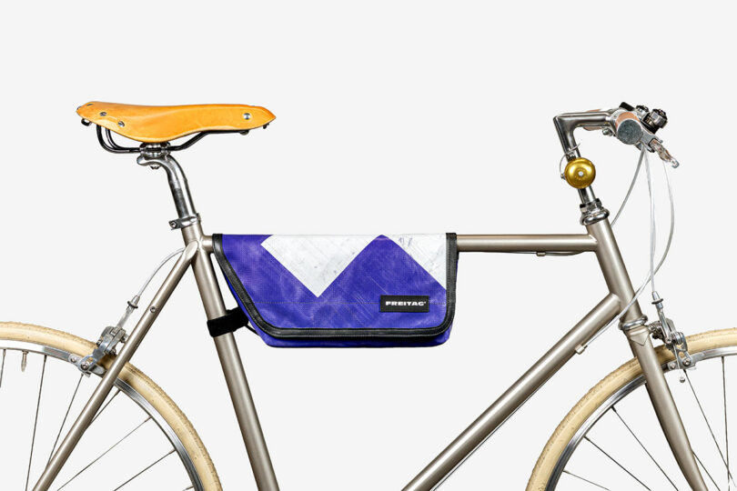 A silver bicycle with tan tires and a tan leather seat, featuring a blue and white FREITAG messenger bag attached to the frame.