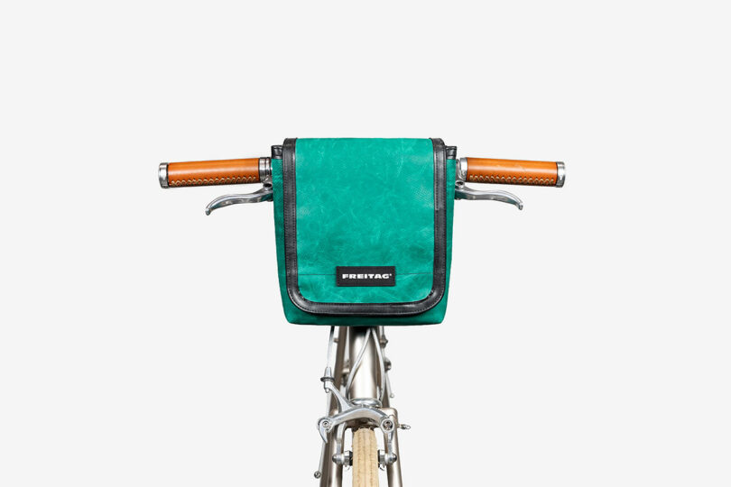 A green Freitag handlebar bag is attached to the handlebars of a bicycle with brown leather grips, photographed against a white background.