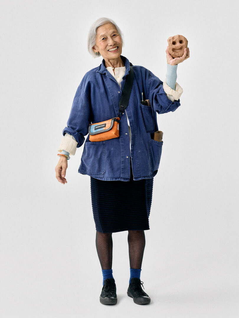 An older woman with gray hair smiles while holding a small, pinkish mask in her raised hand. She wears a blue jacket, black skirt, dark shoes, and a crossbody bag.