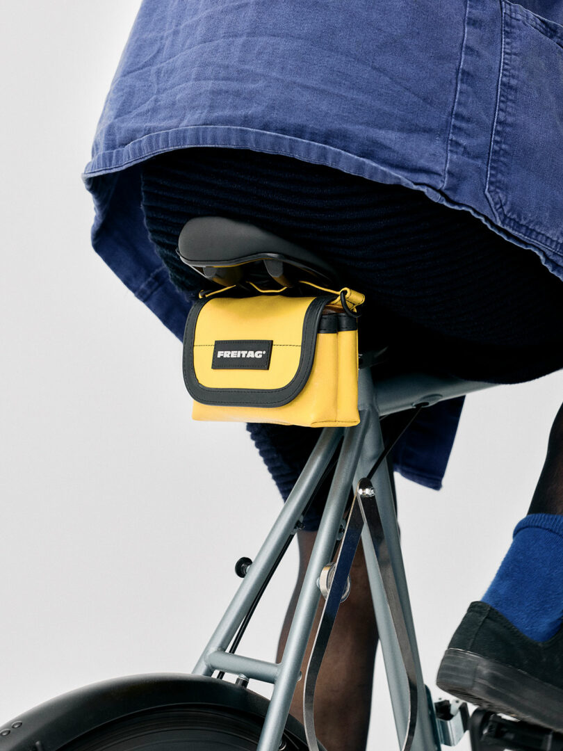Close-up of a person wearing a blue skirt on a bicycle, with a small yellow and black FREITAG bag attached under the seat.
