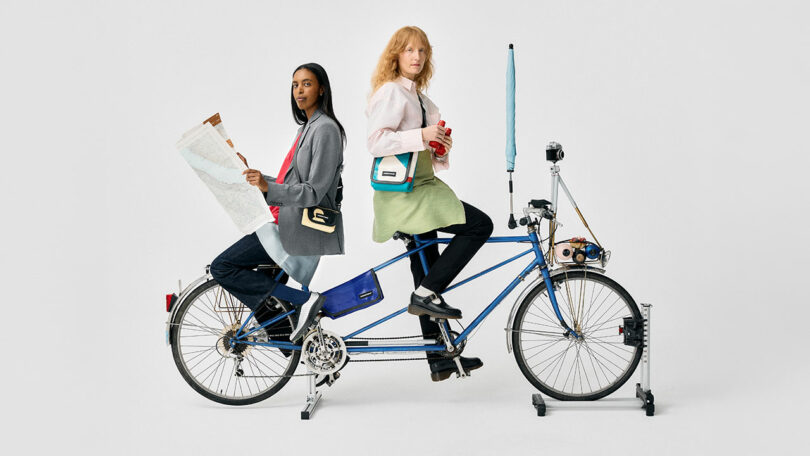 Two women sit on a stationary tandem bicycle; one holds a map, the other a camera, with accessories and bags, against a plain white background.