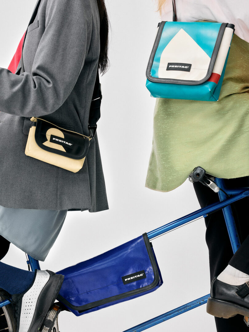 Two people wearing crossbody Freitag bags stand next to a blue bicycle, with a third Freitag bag attached to the bike frame.