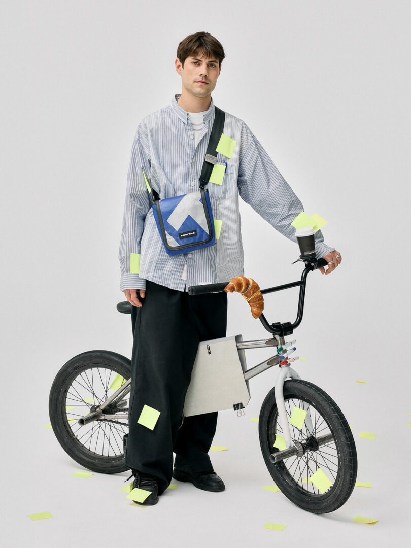 A man stands beside a BMX bike covered in yellow sticky notes. He wears oversized pants, a striped shirt, a shoulder bag, and holds the handlebars with a croissant attached.