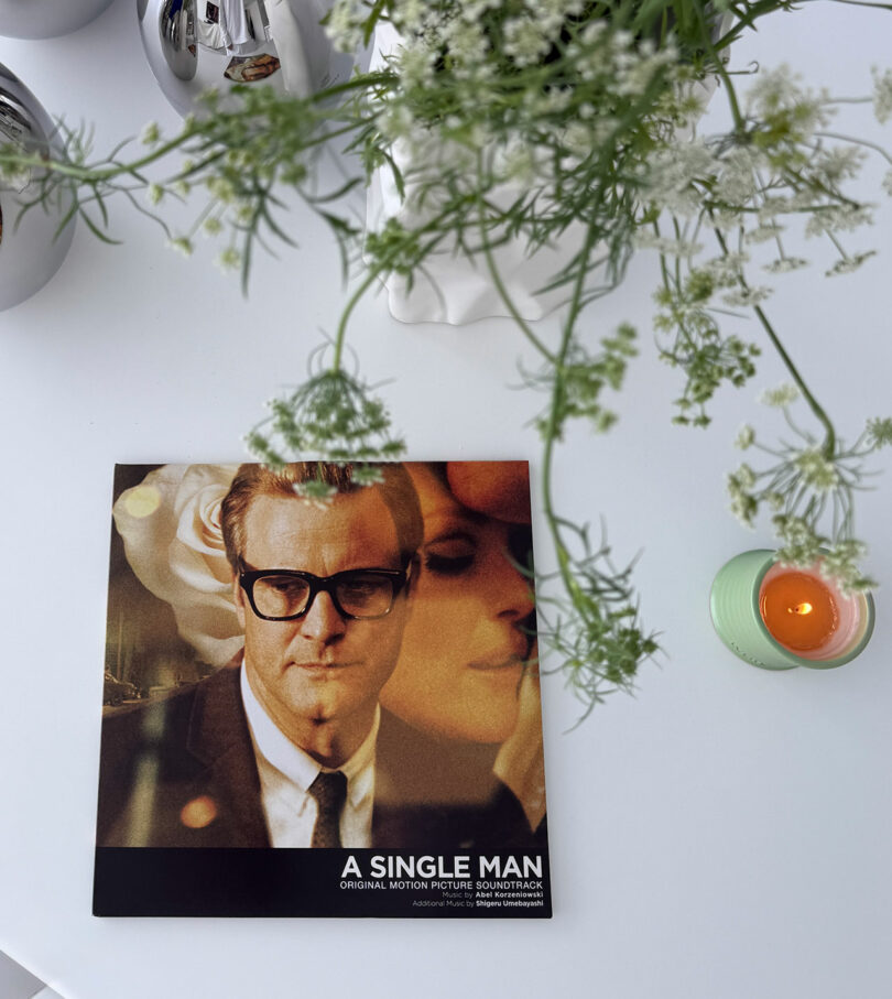 A Kameh vinyl record of "A Single Man" soundtrack lies on a white table next to a green candle and a vase with white flowers.