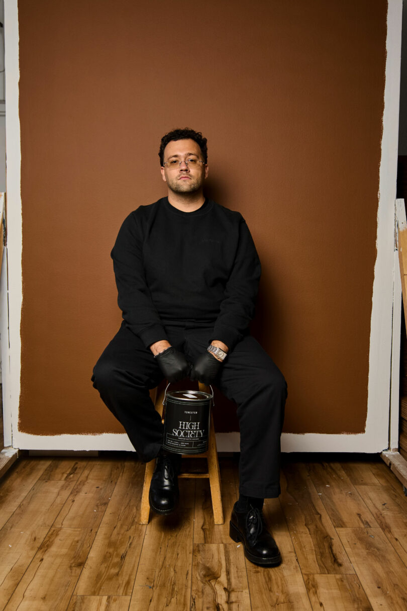 A man in black clothing sits on a wooden stool against a brown backdrop, resting his hands on a paint can labeled "High Society"—a stylish portrait perfect for this week's Friday Five feature.