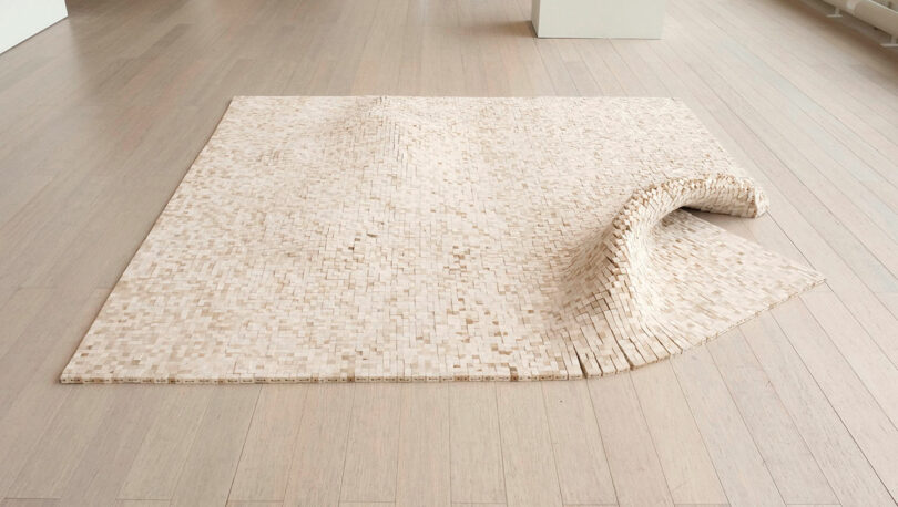 A flat, rectangular wooden tile mosaic by Vivian Chiu lies on a light wooden floor, with one corner artistically raised and bent upward.