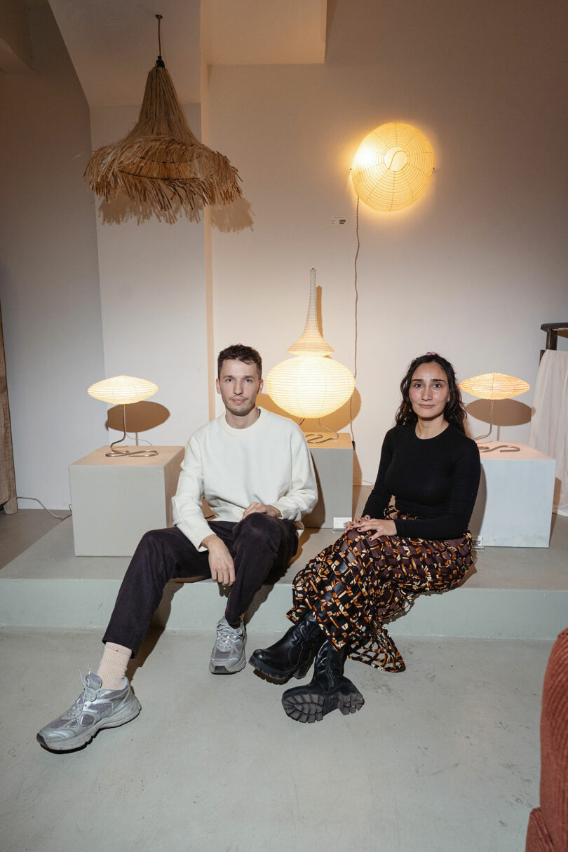 Two people sit indoors on a low platform, surrounded by modern decorative lamps. The man holds a book, and both look toward the camera. The background is softly lit.