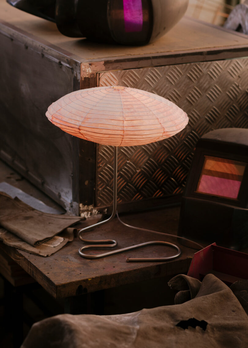 A table lamp with a round, paper-like shade sits on a workbench near welding helmets, gloves, and metal storage boxes.