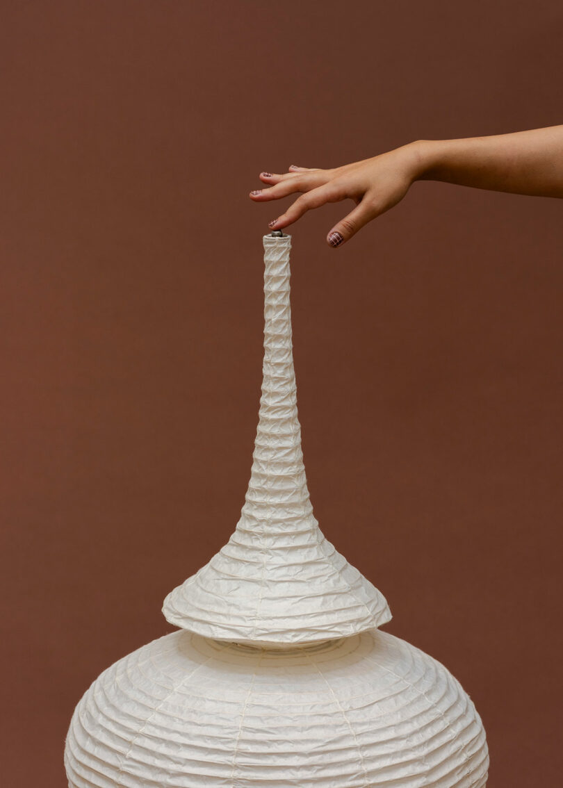 A hand with painted nails reaches toward the top of a tall, white, textured paper sculpture against a plain brown background.