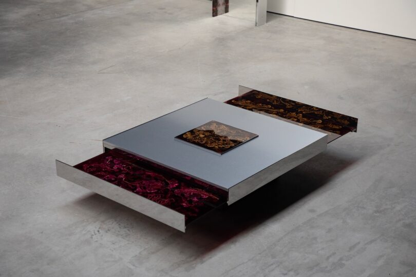A square metallic table with two drawers partially open, revealing marbled red and brown interiors, and a small rectangular object placed on top.