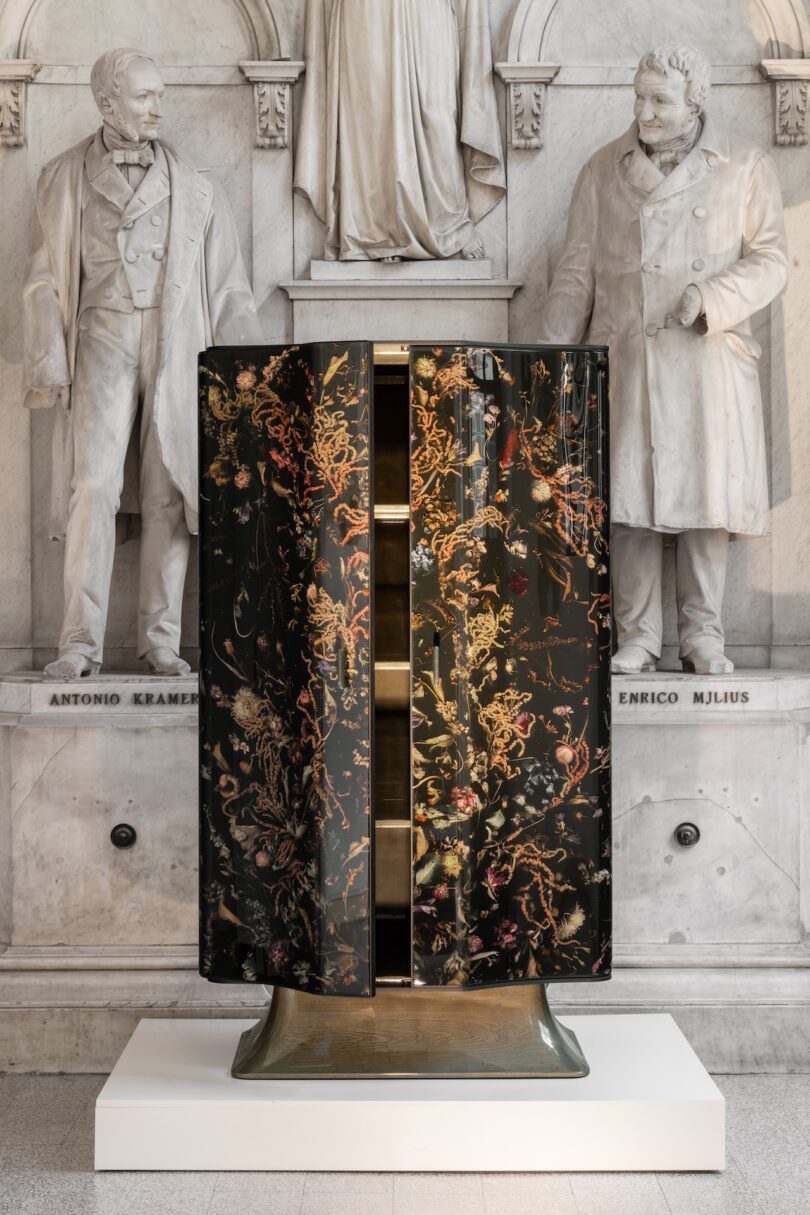 A decorative cabinet with floral patterns stands on a pedestal in front of two white marble statues set in a classical architectural façade.