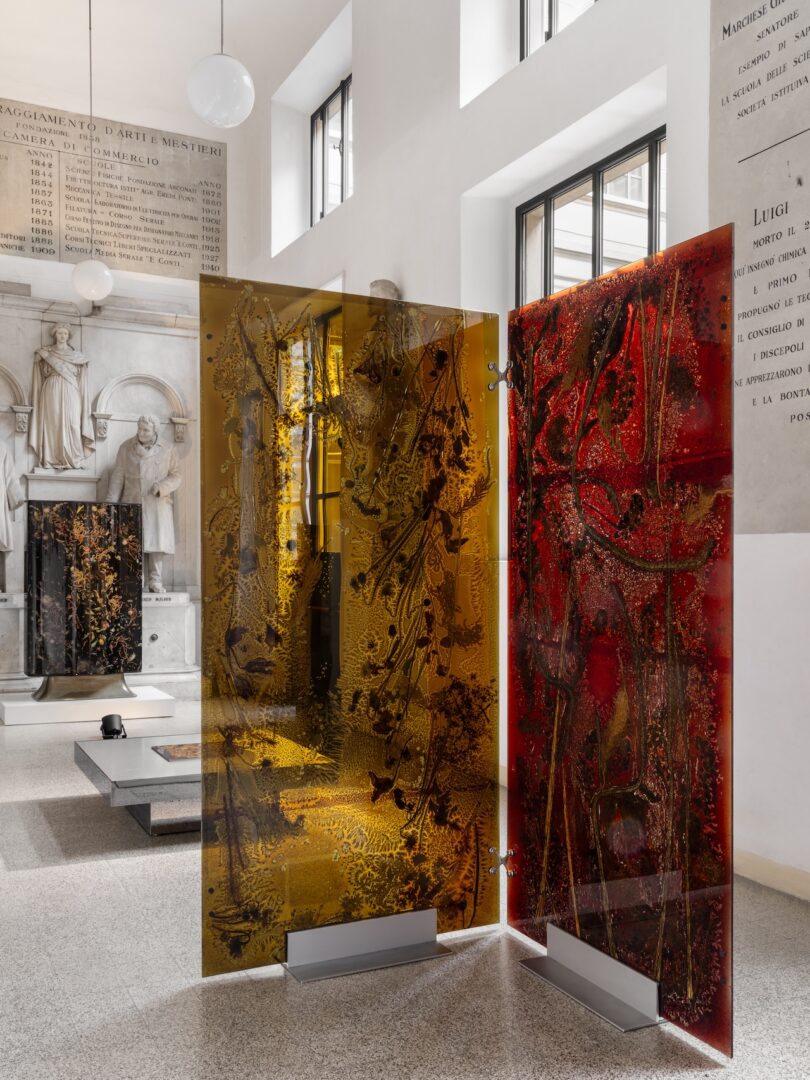 Two large, vertically standing glass panels—one amber, one red—display abstract designs in a museum-like interior with windows and engraved stone walls.