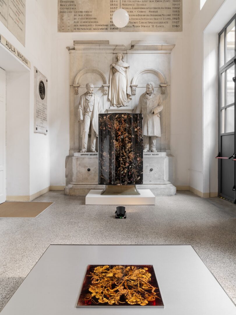 A room with classical statues and plaques, featuring two contemporary art pieces with intricate gold and black designs displayed in the foreground and center.