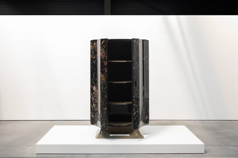 A tall, black, floral-patterned cabinet with open shelves is displayed on a white platform in a minimalist gallery setting.