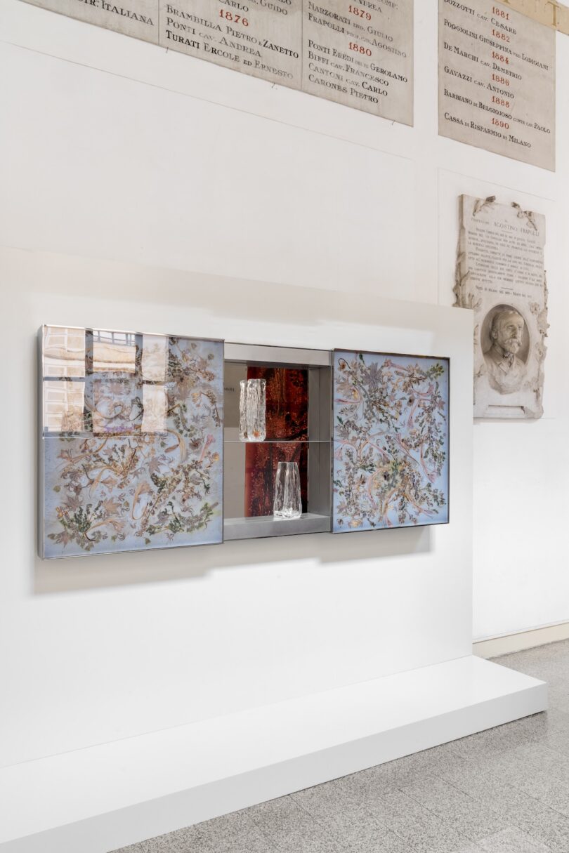 Wall-mounted glass cabinet with mirrored side panels, containing glass objects, displayed on a white platform against a pale wall with engraved plaques and inscriptions.