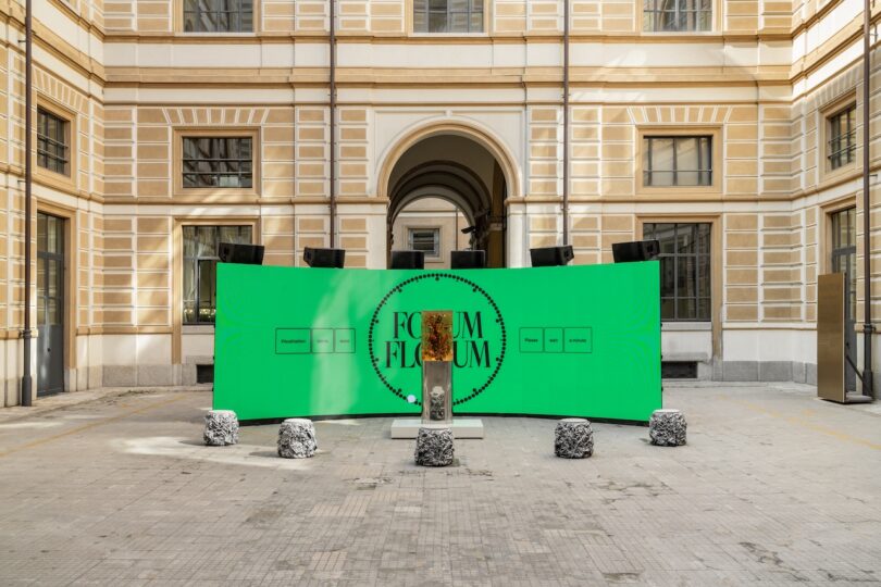 A green digital screen with "FORUM FLORUM" displayed stands in a courtyard; a bust sculpture on a pedestal and four black-and-white patterned stools are arranged in front.
