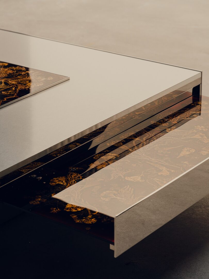 A modern table with a reflective metal surface and transparent sides, revealing a decorative pattern with gold and black designs underneath.