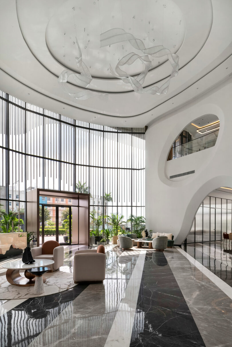 Modern hotel lobby with tall windows, marble floors, white curved walls, contemporary furniture, and indoor plants—a confluence of style—highlighted by a sculptural ceiling light fixture.