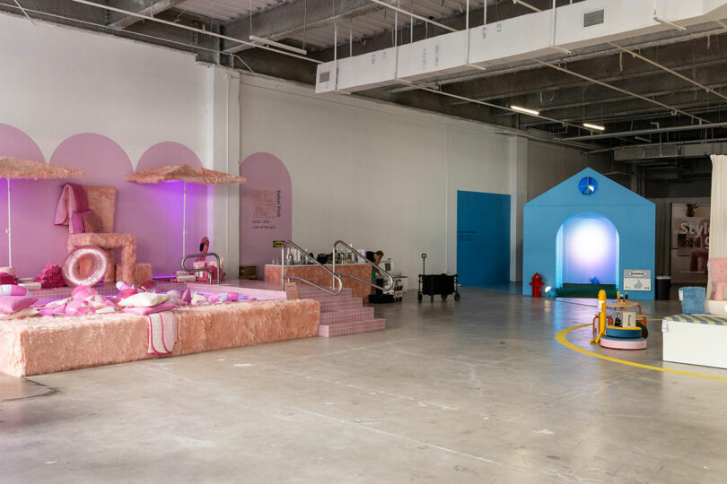 An indoor space features playful, pastel-colored installations, including a pink seating area and a blue house structure, with exposed industrial ceilings above.