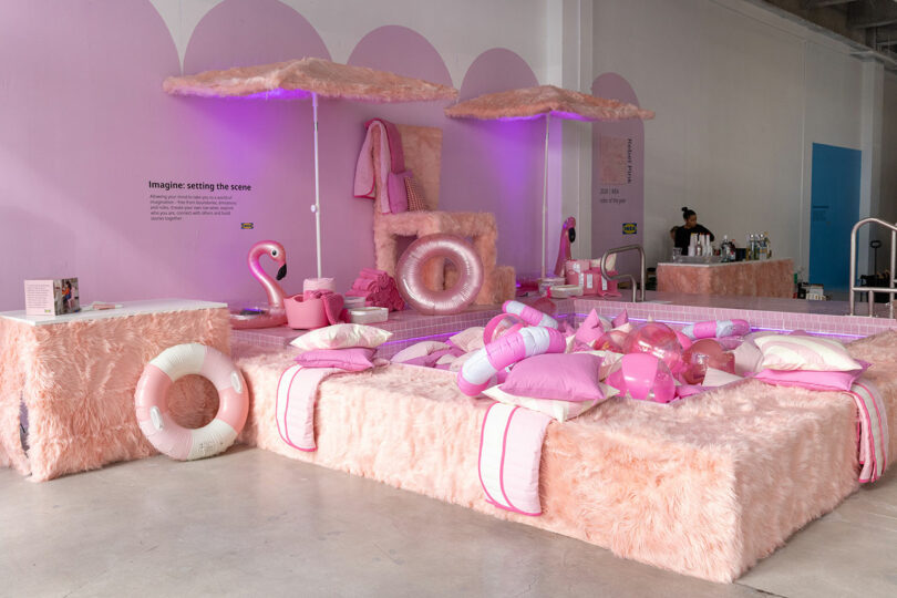 A display area features pink faux fur furniture, inflatable pool floats, pillows, and towels under purple lighting, with a person standing at a bar in the background.
