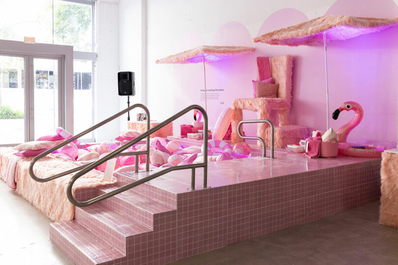 A pink-themed indoor display features faux fur furniture, pink tiled steps, umbrellas, scattered cushions, and a large inflatable flamingo under purple lighting.