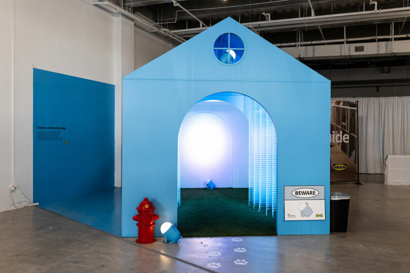 A large blue doghouse structure with an arched entrance, illuminated interior, artificial grass, a red fire hydrant, and a "Beware" sign, displayed indoors on a concrete floor.