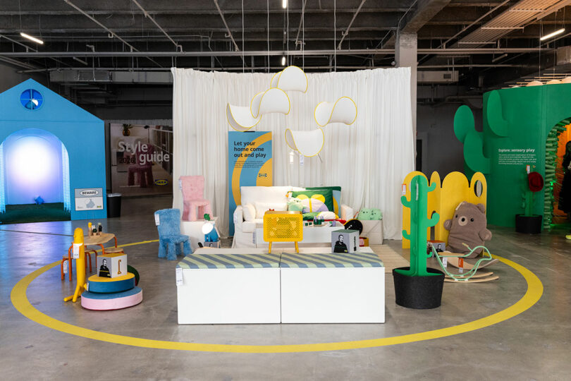 A colorful showroom displays playful, modern furniture and decor with whimsical shapes and bold colors, set on a concrete floor with a white curtain backdrop and bright lighting.