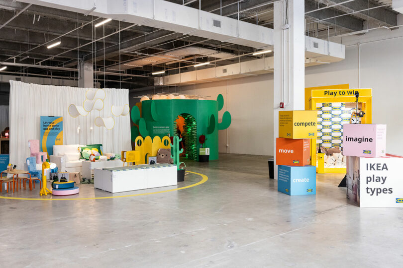 An indoor IKEA exhibit features colorful furniture displays, playful decor, large signage, and interactive zones in a spacious, industrial-style setting.