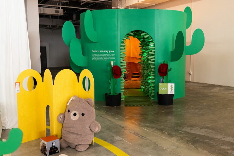 Green cactus-themed sensory play installation with an arched entrance, yellow abstract partition, plush bear toy, and concrete floors in an indoor space.