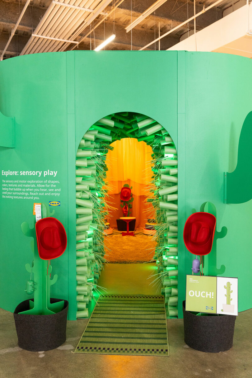 A green sensory play exhibit features an archway lined with soft spikes, red hat-like objects, green decor shaped like cacti, and signage inviting children to explore tactile experiences.