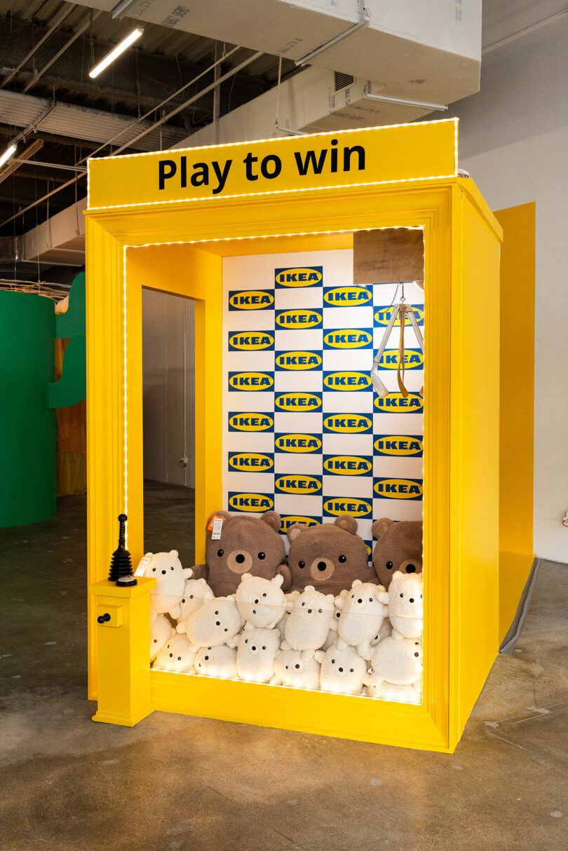 A yellow claw machine booth with "Play to win" sign, IKEA logos inside, and plush bear toys stacked at the front.