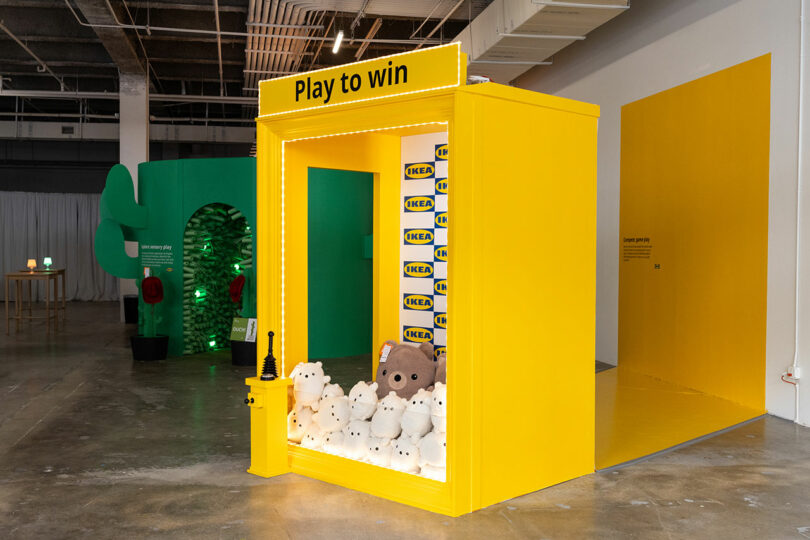 A bright yellow claw machine labeled "Play to win" filled with IKEA plush toys sits in a spacious indoor area with industrial ceilings.