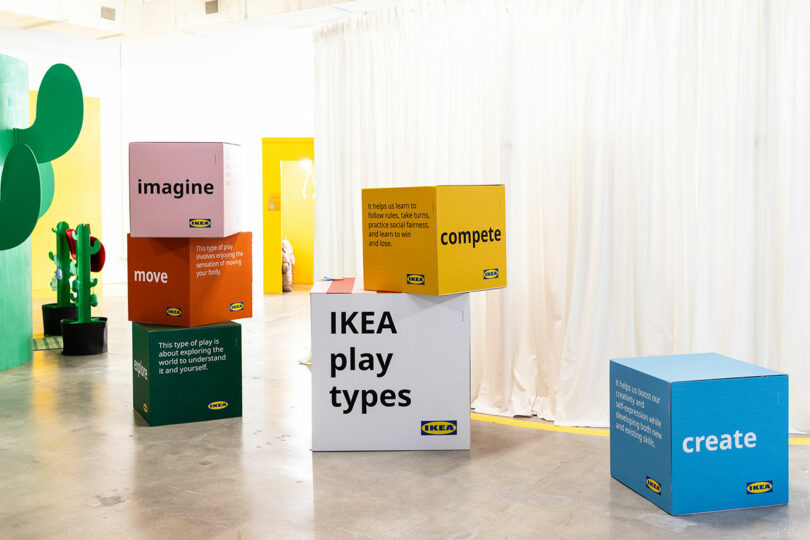 Large colored cubes with motivational words like "imagine," "move," "compete," and "create" are displayed in a modern, minimal indoor space with a white curtain and colorful decor.