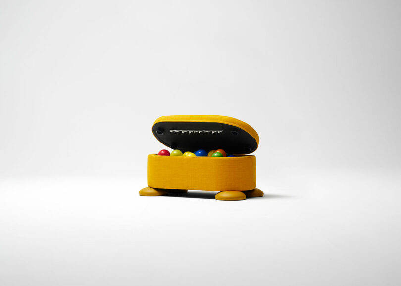 A small, yellow, box-shaped container with a hinged lid, resembling a creature with eyes and teeth, holds colorful plastic balls inside.