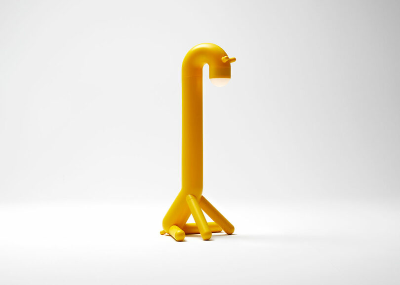 A yellow, dog-shaped lamp with a rounded body, four short legs, and a light source at the end of its curved neck, set against a plain white background.