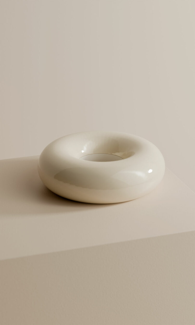 A glossy, white, donut-shaped ceramic object sits on a light beige surface against a neutral background.