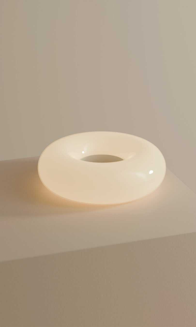 A round, smooth, white, donut-shaped light sits on a beige pedestal against a plain background.