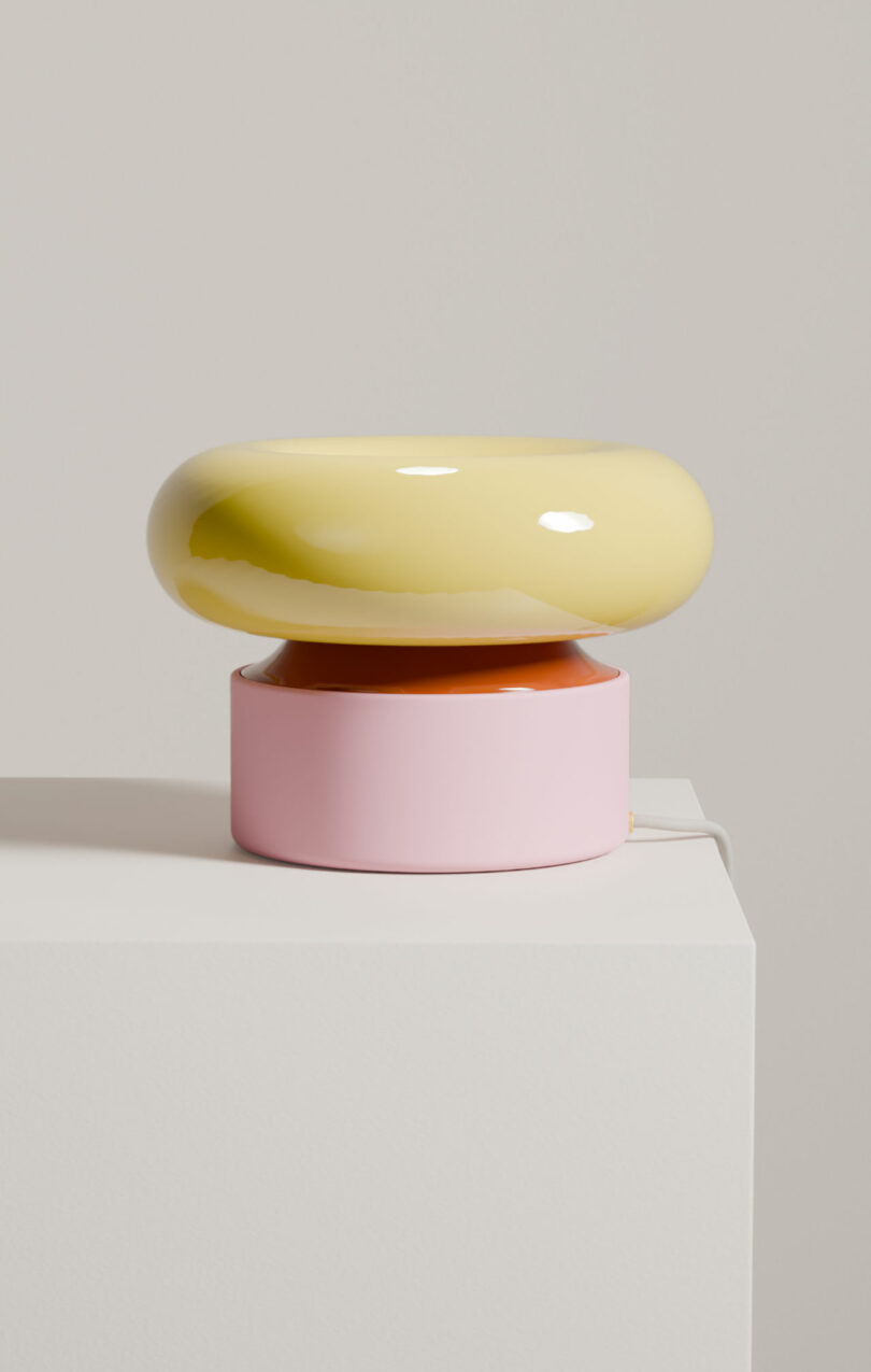 A modern table lamp with a glossy yellow, rounded top and a cylindrical pink base sits on a white pedestal against a neutral background.