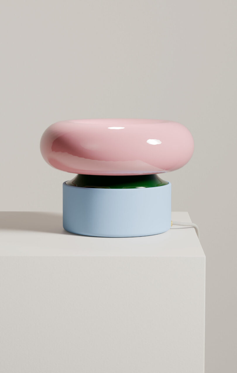 A modern table lamp with a glossy pink rounded top, green middle section, and light blue cylindrical base, sitting on a white surface against a plain background.