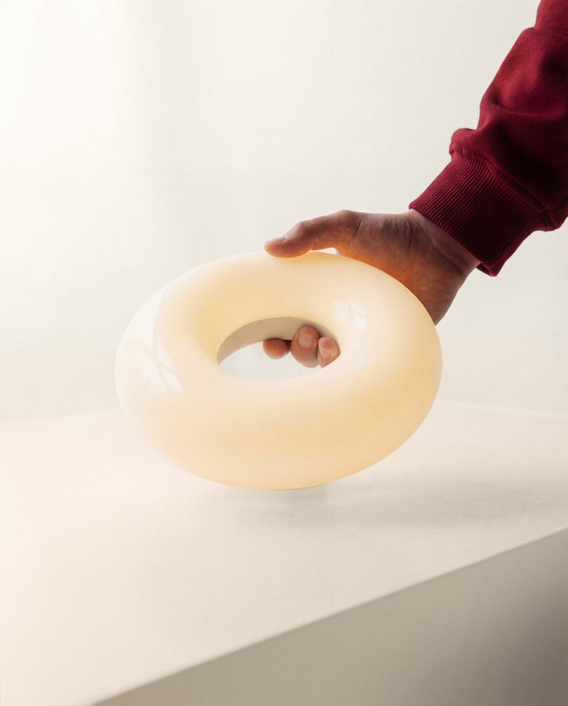 A hand in a red sleeve holds a glowing, round, donut-shaped lamp on a white surface.