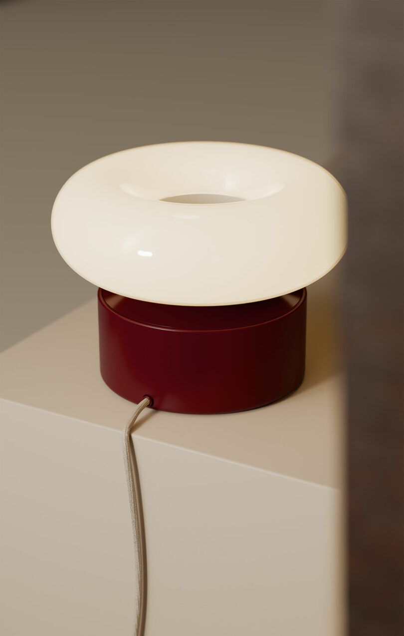 A modern table lamp with a white circular top and a maroon cylindrical base, placed on a beige surface with a cord extending from the base.