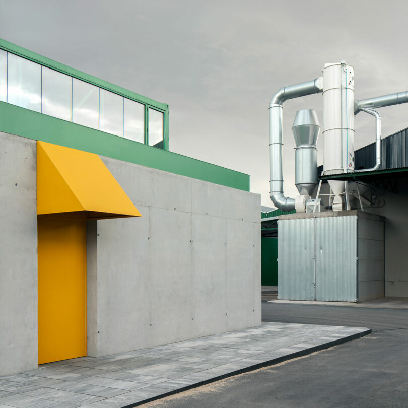 A modern industrial building, possibly an Impepinable Studio, with a gray concrete wall, a bright yellow door canopy, metal ventilation pipes, and green accents under a cloudy sky.
