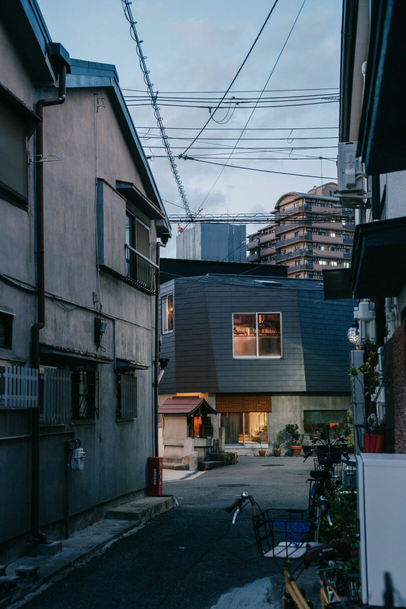 A narrow street lined with residential buildings, bicycles, and overhead wires, with a modern house and high-rise buildings visible in the background.